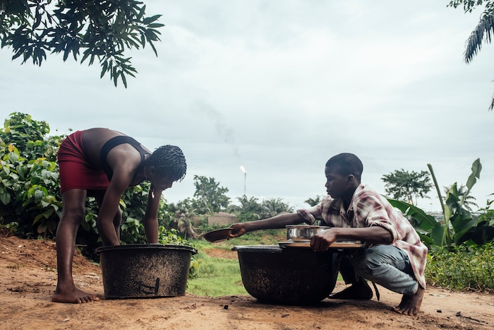 (KC Nwakalor | The New York Times) Ada Osi 16 and Promise Osi, 14, help with the dishes on their family's cassava farm in Obrikom, in the heart of Nigeria's oil-rich delta, July 20, 2020. Around the world, the poor and marginalized are much more likely to be vulnerable to extreme heat; methane gas flares burning around the clock in Obrikom make this already hot area worse still.