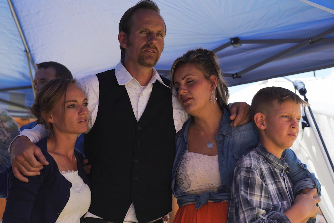 (Shannon Mullane for High Country News) Enoch Foster embraces his third wife, Lydia, left, and his second wife, Lillian, right, as she holds her son Elijah, during a closing hymn and prayer at the testimony meeting at Rockland Ranch on July 28, 2019.