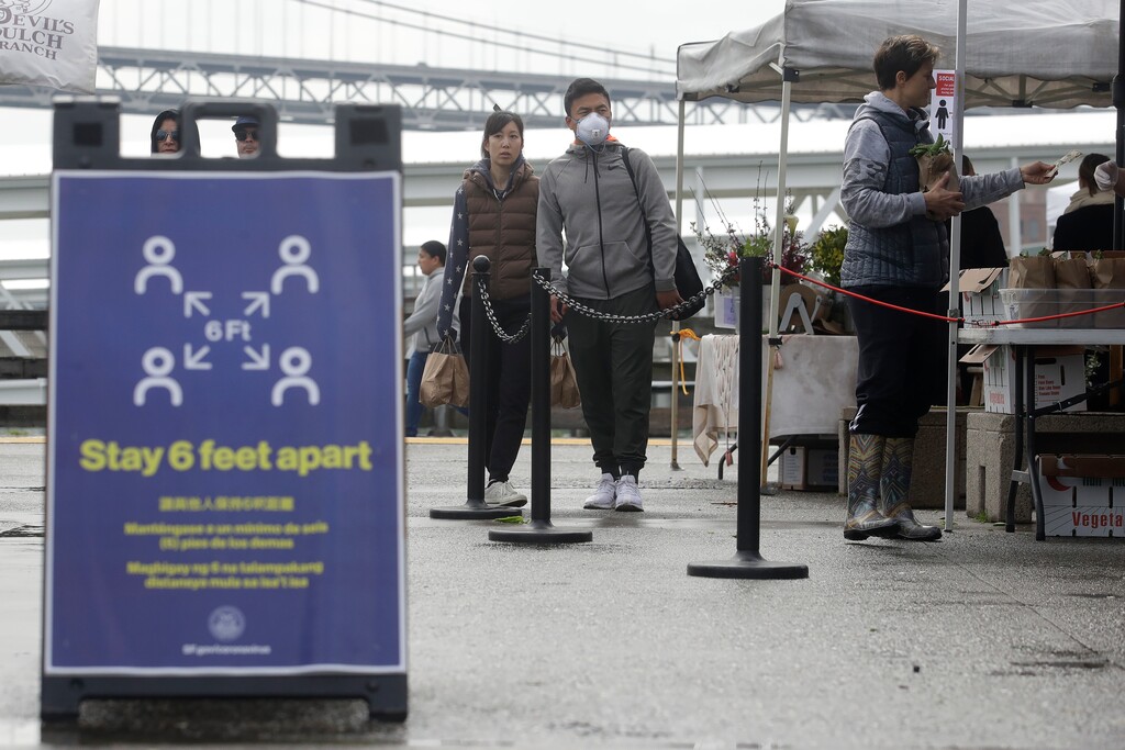 (Jeff Chiu | AP) A sign is shown advising people to maintain distance is shown as a man wears a mask while waiting in line at the Ferry Plaza Farmers Market in San Francisco, Saturday, March 28, 2020.