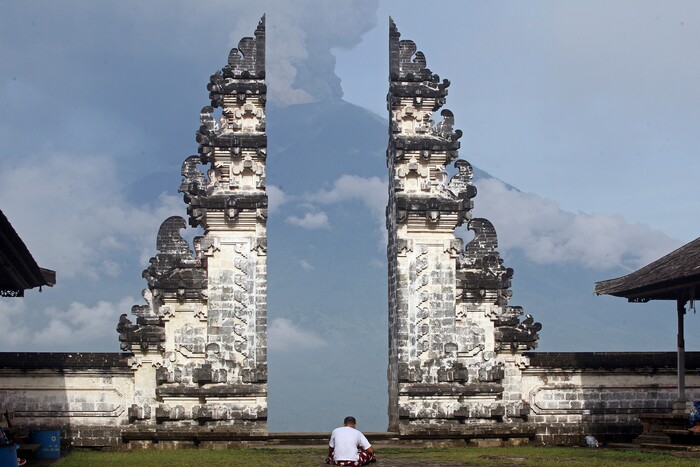 (Firdia Lisnawati | The Associated Press) A man sits at a temple with the Mount Agung volcano erupting in the background in Karangasem, Indonesia, Monday, Nov. 27, 2017. Indonesia authorities raised the alert for the rumbling volcano to highest level on Monday and closed the international airport on tourist island of Bali stranding thousands of travelers.