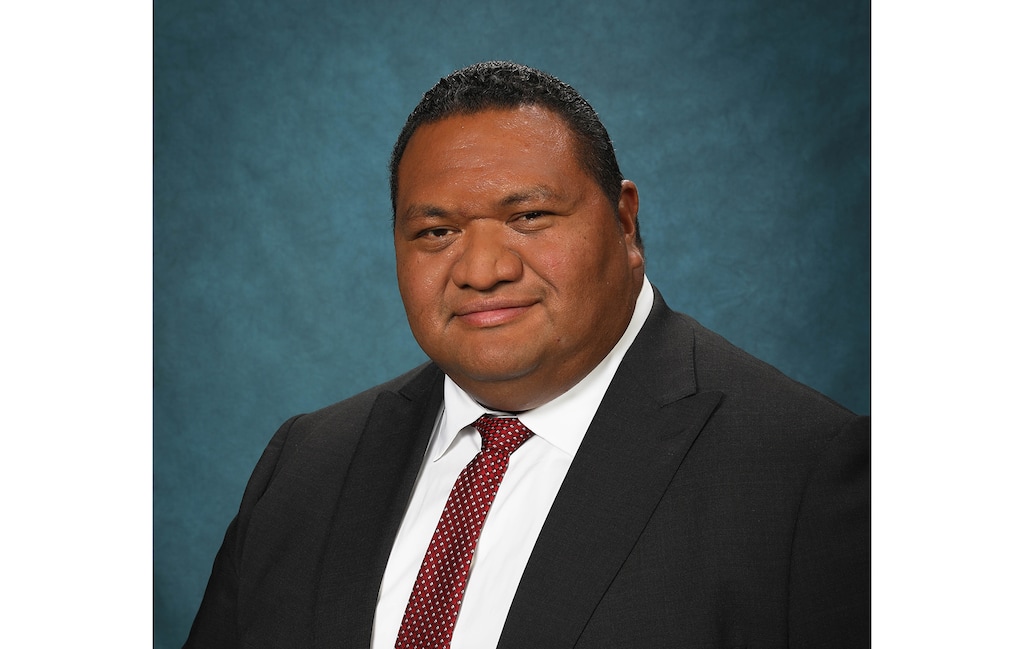 A man in a dark suit jacket, white dress shirt and red tie looks at the camera in front of a blue background.