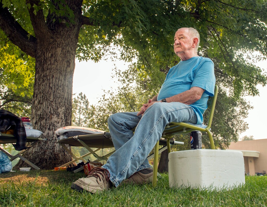 (Hector Amezcua | The Sacramento Bee | The Associated Press) Otis Bershers, 75, of the city of Shasta Lake, sits in a chair at the Shasta College evacuation center, on Sunday, July 29, 2018, in Redding, Calif., were he has slept outside with his wife, Carol, and cat Smokey since Thursday.