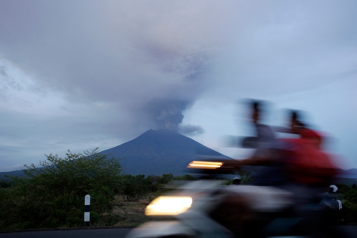 (Firdia Lisnawati | The Associated Press) A motorcyclist passes by the Mount Agung volcano erupting in the background in Karangasem, Indonesia, Monday, Nov. 27, 2017. Indonesia authorities raised the alert for the rumbling volcano to highest level on Monday and closed the international airport on tourist island of Bali stranding thousands of travelers.