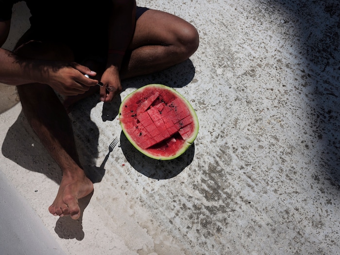 (Myrto Papadopoulos | The New York Times) Hasib Hotak, a homeless refugee from Afghanistan, eats watermelon outside the rooftop shack where he lives in Athens, Greece, July 22, 2020. Around the world, the poor and marginalized are much more likely to be vulnerable to extreme heat; Hasib sleeps with other refugees on a rooftop that turns blazing hot by midday.