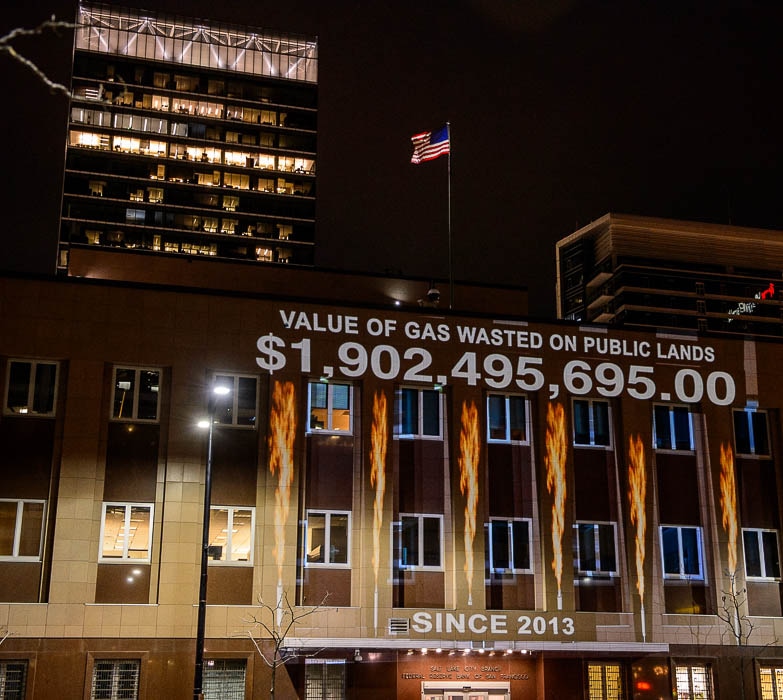 (Trent Nelson | The Salt Lake Tribune)  In a visual protest, the group Alliance for Better Utah projects the total dollar value of natural gas lost on public lands since 2013 due to venting, flaring, and leaking onto the side of Federal Reserve Building in Salt Lake City, Thursday April 5, 2018. President Trump’s Interior Department is poised to rollback Obama-era rules requiring oil and gas operations on public lands to capture that natural gas instead of burning it.