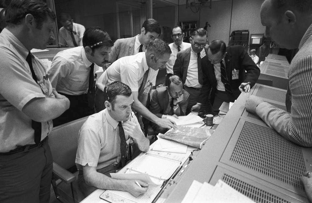 (NASA | AP) In this April 15, 1970 photo made available by NASA, a group of flight controllers gather around the console of Glenn S. Lunney, foreground seated, Shift 4 flight director, in the Mission Operations Control Room (MOCR) of Mission Control Center (MCC) in Houston. Their attention is drawn to a weather map of the proposed landing site in the Pacific Ocean. At this point, the Apollo 13 lunar landing mission had been canceled, and the problem-plagued Apollo 13 crew members were in trans-Earth trajectory attempting to bring their crippled spacecraft back home.
