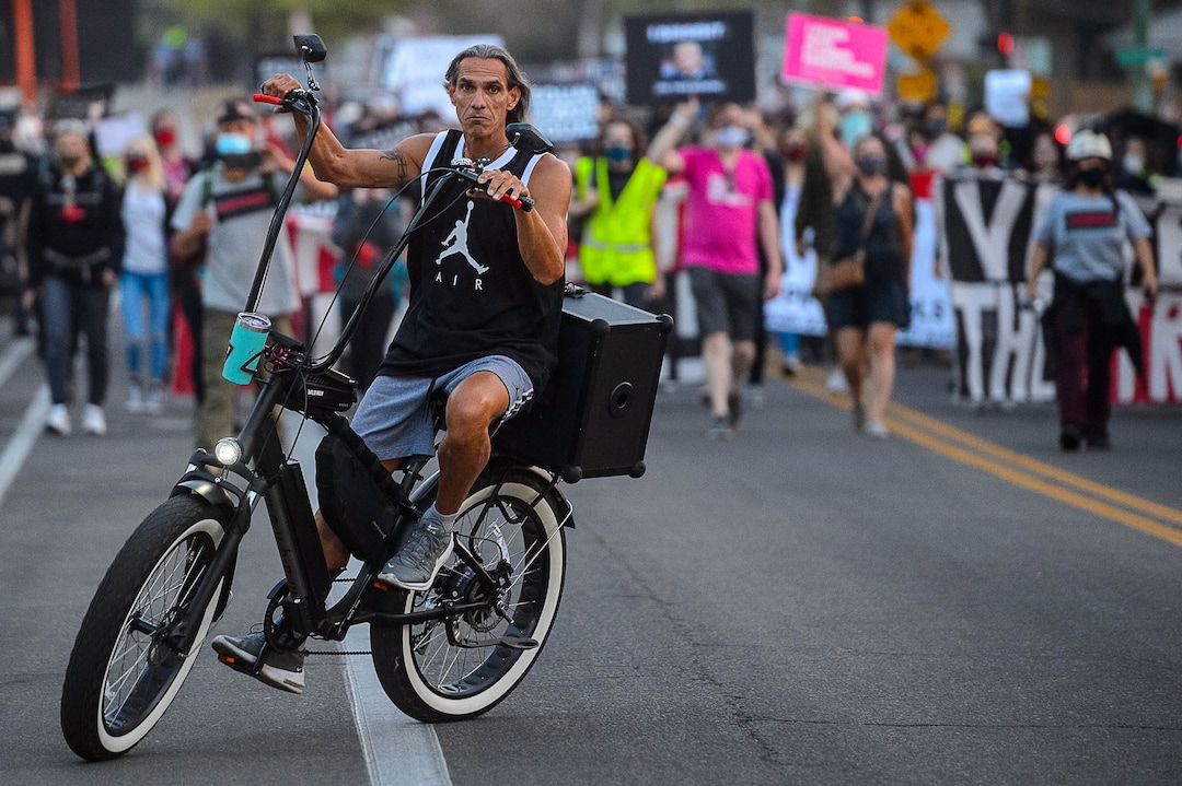 (Trent Nelson  |  The Salt Lake Tribune) A man rides a bike while blasting the song "Proud to be an American" near protesters marching before Vice President Mike Pence and Sen. Kamala Harris were to debate at Kingsbury Hall in Salt Lake City on Wednesday, Oct. 7, 2020.