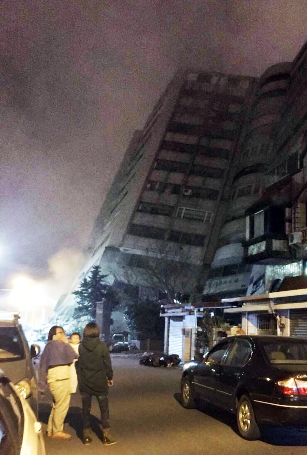 In this photo released by Hualien County Fire Bureau, Taiwanese people wath a building that collapsed on its side from an early morning earthquake in Hualien County, eastern Taiwan, early Wednesday, Feb. 7 2018.  A 6.4-magnitude earthquake has struck eastern Taiwan, according to the U.S. Geological Survey. (Hualien County Fire Bureau via AP)