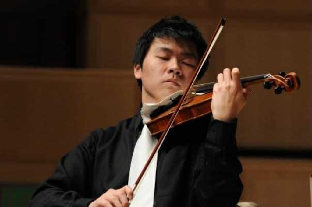 A man in a black shirt and white tie closes his eyes while he plays a violin.