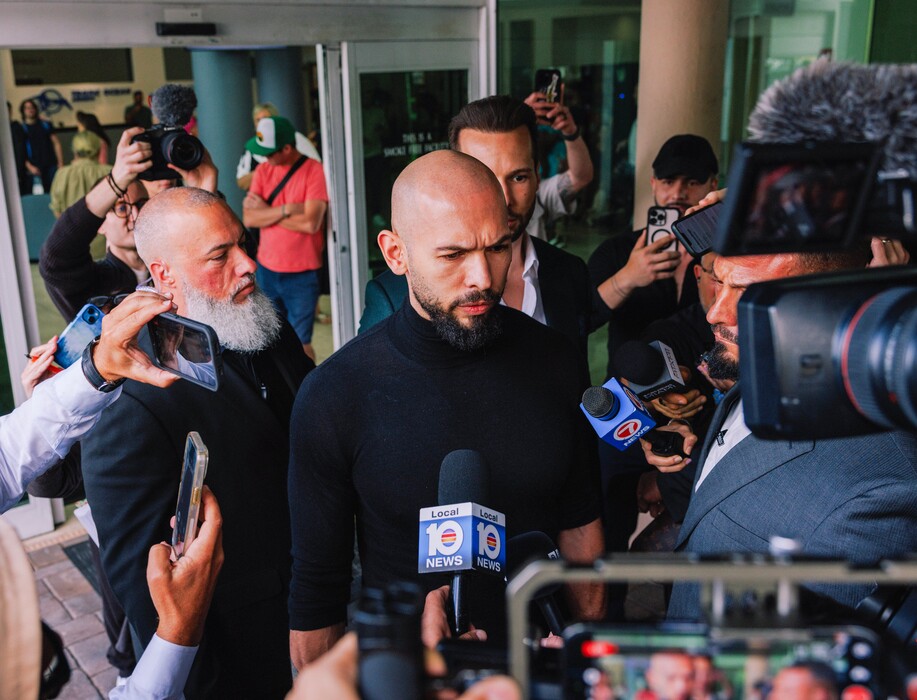 (James Jackman | The New York Times) The British American online influencer Andrew Tate speaks to reporters after he and his brother, Tristan Tate, obscured at center right, arrived at Fort Lauderdale-Hollywood International Airport in Florida on Thursday, Feb. 27, 2025. The brothers, who had been barred from leaving Romania for more than two years over criminal investigations, were allowed by a judge to leave on Thursday, a development that raised questions about whether the Trump administration played a role in their sudden departure.