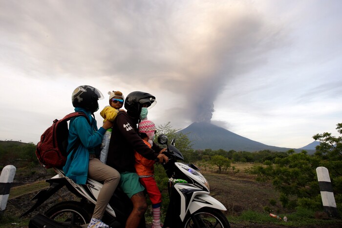 (Firdia Lisnawati | The Associated Press) A family on a motorcycle passes by the Mount Agung volcano erupting in the background in Karangasem, Indonesia, Monday, Nov. 27, 2017. Indonesia authorities raised the alert for the rumbling volcano to highest level on Monday and closed the international airport on tourist island of Bali stranding thousands of travelers.
