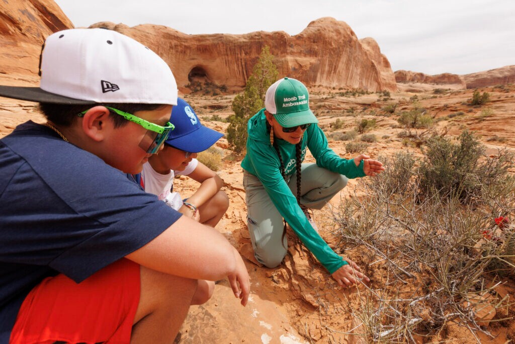 (Grand County Active Transportation and Trails via Corie Spruill) A Moab Trail Ambassador explaining the function of biological soil crust to kids on Corona Arch.