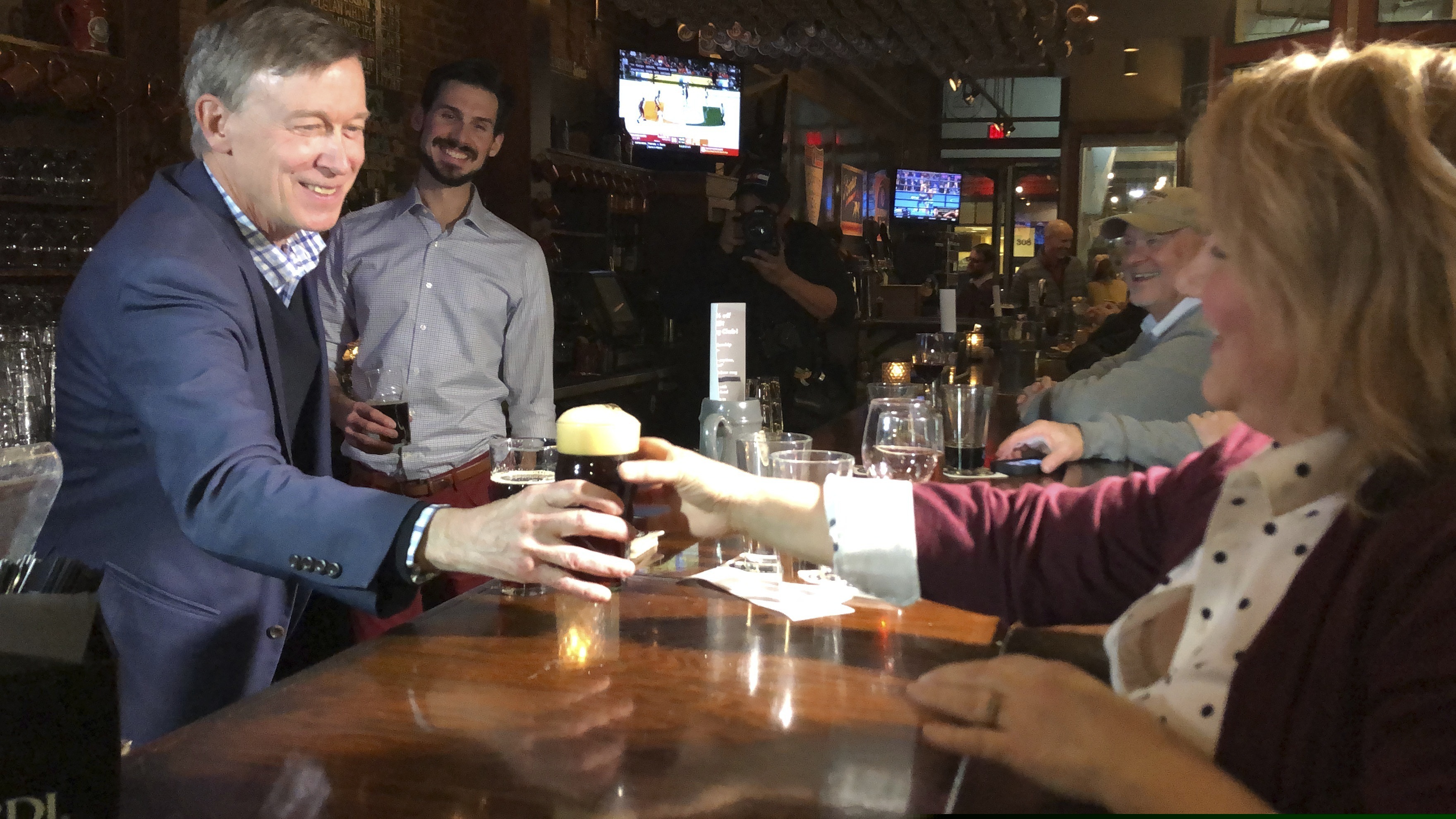 In this Jan. 27, 2019, photo, former Colorado Gov. John Hickenlooper, left, hands a patron a beer during a stop at Court Avenue Brewing Co. in Des Moines, Iowa. As the first wave of Democratic presidential candidates unveil plans for taxing wealth and universal government-provided health care, Hickenlooper is making a narrower pitch of beer and bipartisanship. (Brianne Pfannenstiel/The Des Moines Register via AP)