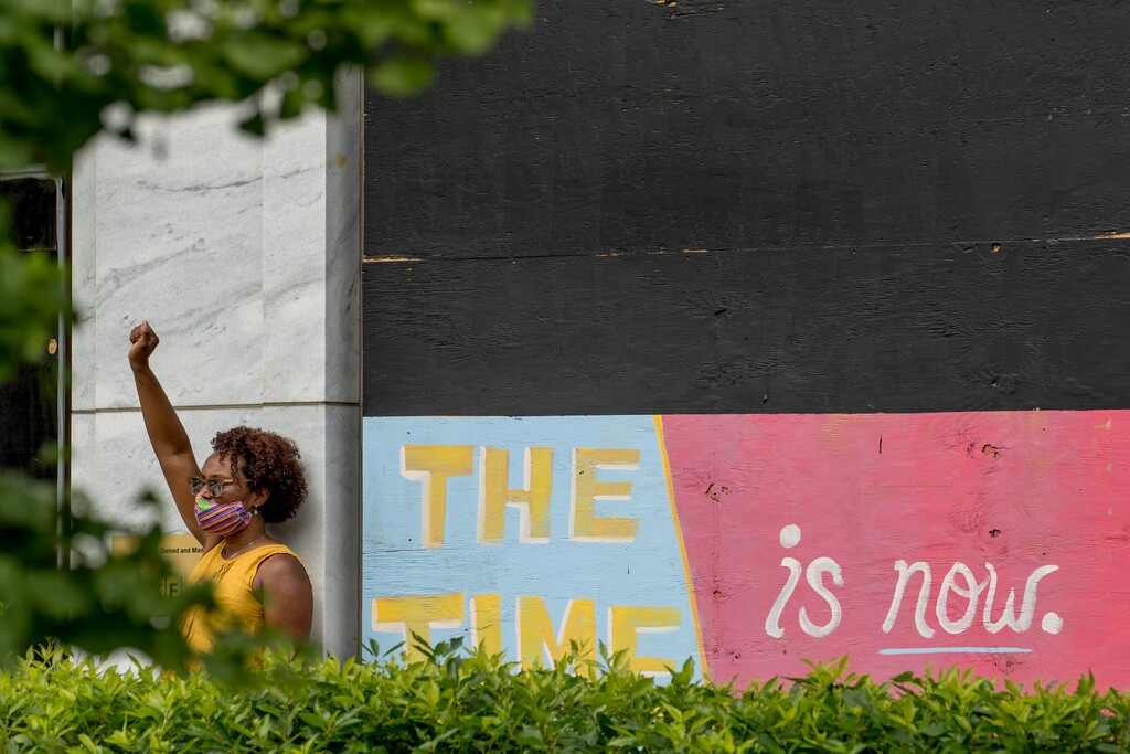 A woman holds her hand up in a fists as she poses for a photograph along with a mural that reads "The Time is Now" along 16th Street Northwest renamed Black Lives Matter Plaza near the White House, Friday, June 19, 2020, in Washington. (AP Photo/Andrew Harnik)