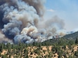 Grey and black smoke clouds billow above a forest of ponderosas and piñon-junipers.