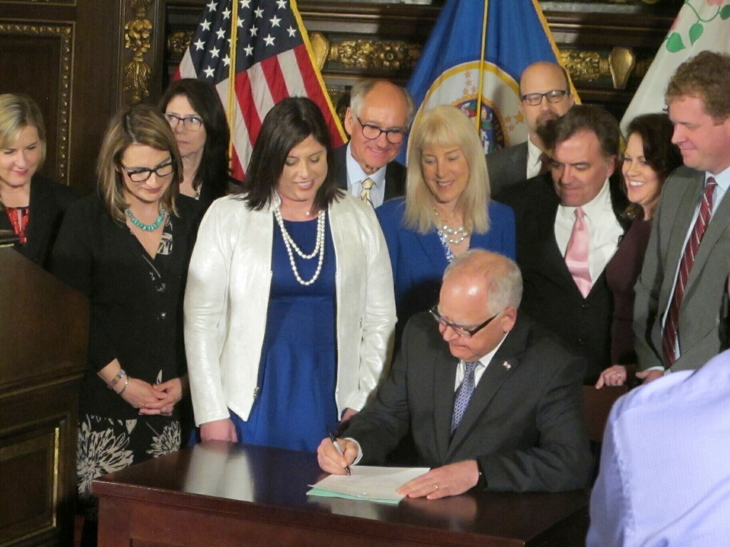 (AP Photo/Steve Karnowski) Jenny Teeson, center in white, of Andover, Minnesota, looks on as Minnesota Gov. Tim Walz signs a bill at the Capitol in St. Paul, on Thursday, May 2, 2019, repealing a Minnesota law that prevented prosecutors from filing sexual assault charges against people accused of raping their spouse. Teeson, testified before legislative committees earlier this year about how her now ex-husband drugged her and made a video of himself raping her while she was unconscious. Prosecutors dropped rape charges because of the old law, and he served just 30 days in jail for invasion of privacy.