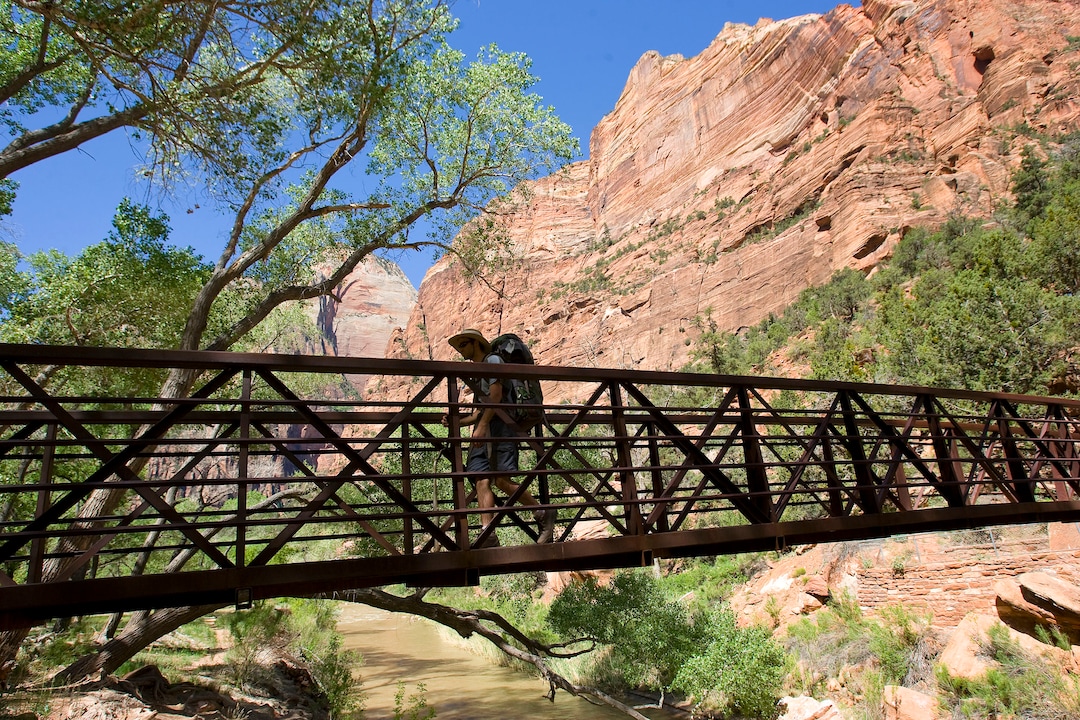 (Al Hartmann | The Salt Lake Tribune) Hiker crosses Virgin River on the green valley floor with surrounding sandstone formations in Zion National Park.