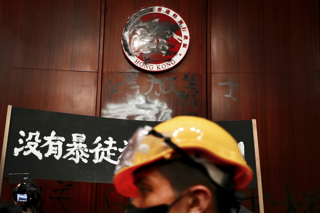(Vincent Yu | AP Photo) A protester walks by a defaced Hong Kong emblem and a banner reads "No thug, only tyranny" after they broke into the Legislative Council building in Hong Kong, Monday, July 1, 2019. Protesters in Hong Kong took over the legislature's main building Monday night, tearing down portraits of legislative leaders and spray painting pro-democracy slogans on the walls of the main chamber.