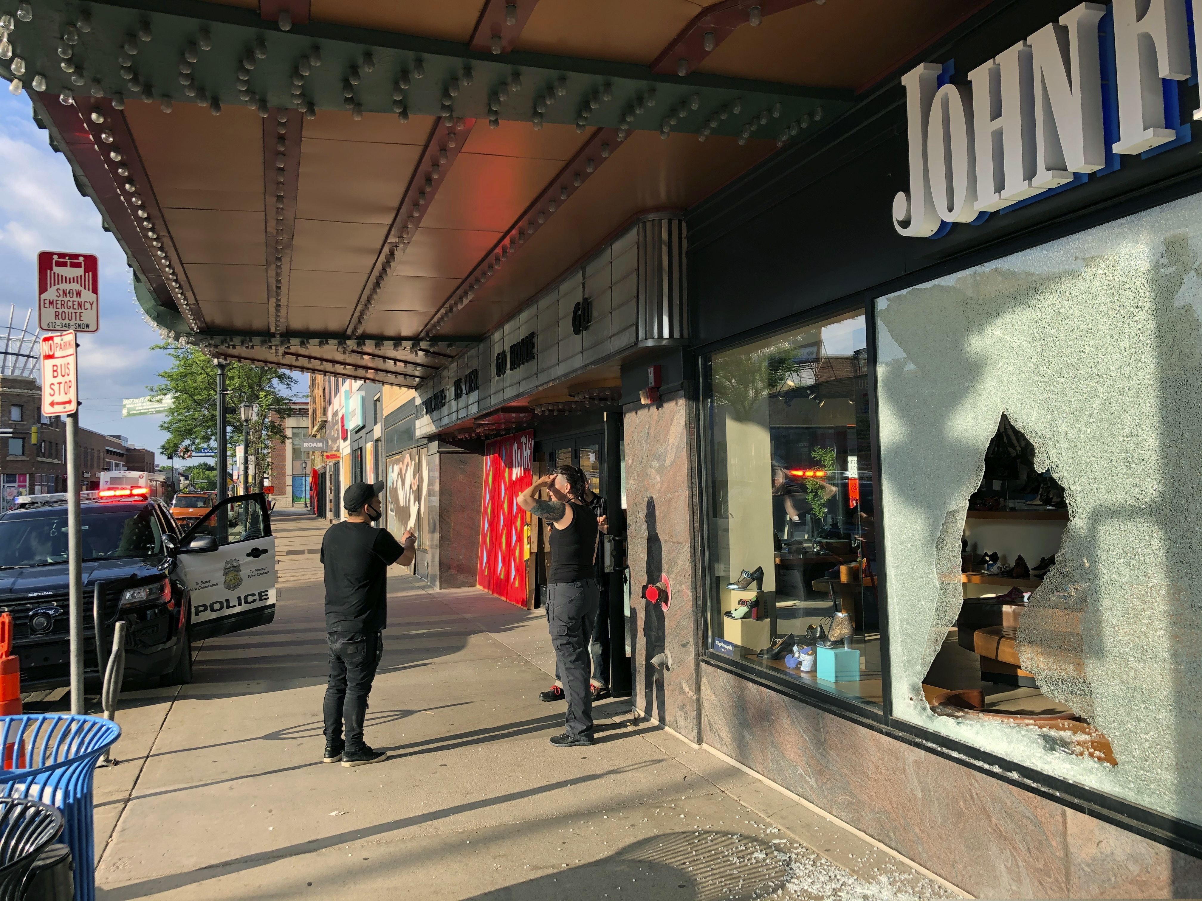 (Doug Glass | The Associated Press) People stand near a storefront of a shoe store damaged by gunfire as a police vehicle remains on the scene outside the Uptown Theatre Sunday, June 21, 2020, following a shooting in Minneapolis' Uptown neighborhood. Multiple people were shot, one fatally, when gunfire broke out shortly after midnight Sunday.