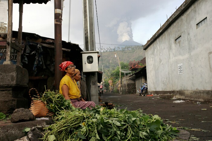 (Firdia Lisnawati | The Associated Press) Vegetables sellers wait for customers at a market with a backdrop of the Mount Agung volcano erupting in Karangasem, Indonesia, Monday, Nov. 27, 2017. Indonesia authorities raised the alert for the rumbling volcano to highest level on Monday and closed the international airport on tourist island of Bali stranding thousands of travelers.