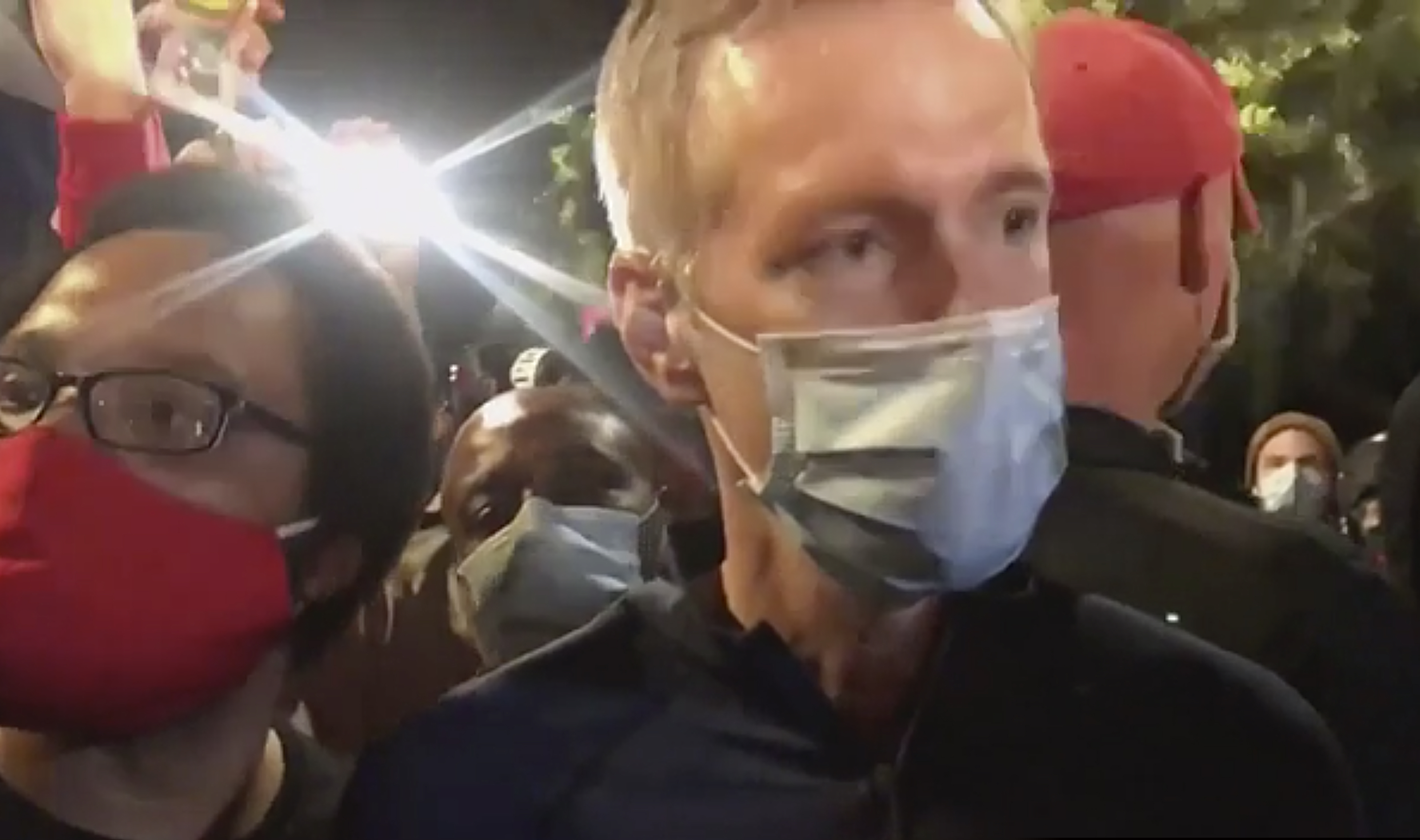 (Karina Brown via AP) In this image made from video released by Karina Brown, Portland Mayor Ted Wheeler, right, stands at a fence guarding a federal courthouse as tear gas drifts by early July 23, 2020, in Portland Oregon, during another night of protest against the presence of federal agents sent by President Donald Trump to quell unrest in the city.
