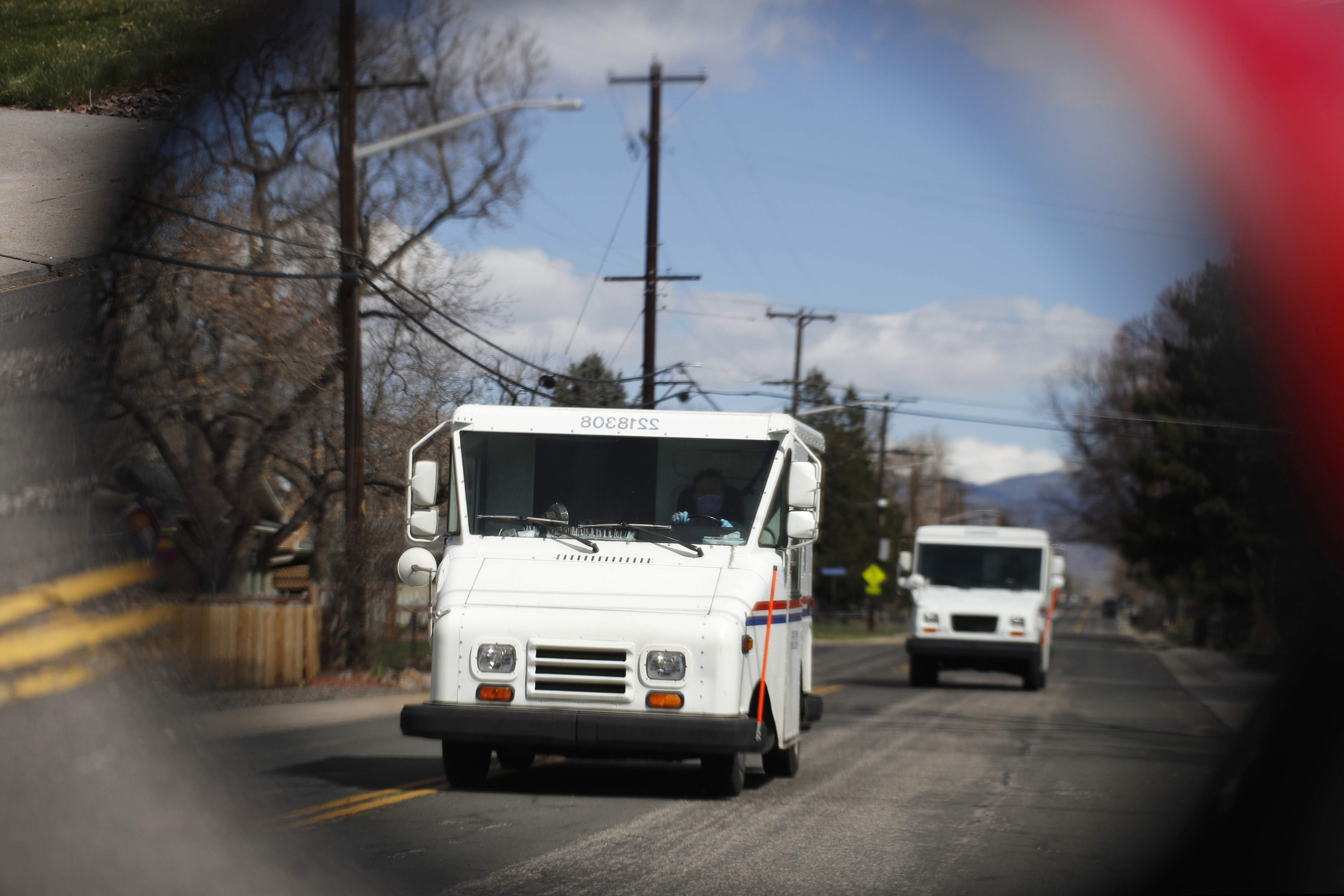 (David Zalubowski | AP file photo)  United States Post Office delivery trucks are reflected in the side mirror of a vehicle as postal delivers set off on their daily rounds in Arvada, Colo., on March 31, 2020.
