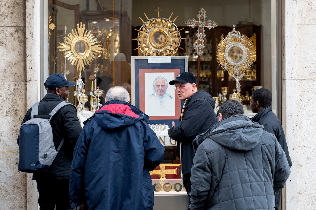 (James Hill | The New York Times) Priests look at a portrait of Pope Francis, with a message asking for people to pray for him, in the window of a shop for religious items near the Vatican in Rome, on Tuesday, Feb. 25, 2025.