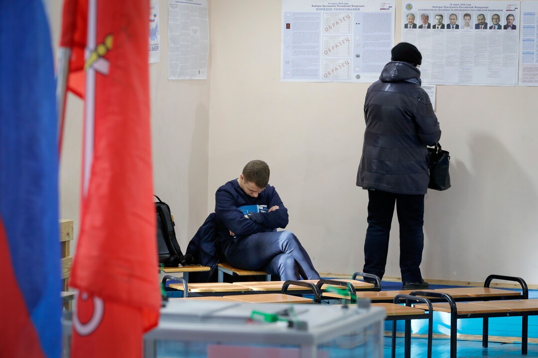 A woman examines a poster of candidates as an observer sleeps at a polling station during presidential elections in St.Petersburg, Russia, Sunday, March 18, 2018. Russians are voting in a presidential election in which Vladimir Putin is seeking a fourth term in the Kremlin. (AP Photo/Dmitri Lovetsky)