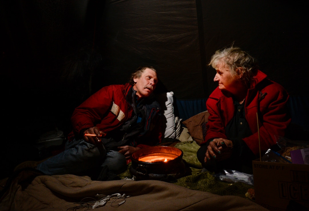 (Leah Hogsten  |  The Salt Lake Tribune)  Ron Barrett, 55, and his wife Katherine Barrett, 54, who are homeless try to stay warm in their tent from the meager heat from a ceramic pot filled with candle wax. Their cold hands are blacken by the soot rising from the burning candle on a night that temperatures dropped to 12 degrees in Salt Lake City. "It's crazy, you gotta be able to have a sense of humor," said Ron Barrett, 55, of the life circumstances that resulted in life on the streets for he and Katherine since 2017. The Barretts are trying to find housing through a Shelter Plus Care housing voucher through the Salt Lake City Housing Authority. The couple says they refuse to stay in the state's largest homeless shelter, The Road Home on Rio Grande Street, because of past threats of violence from other residents.