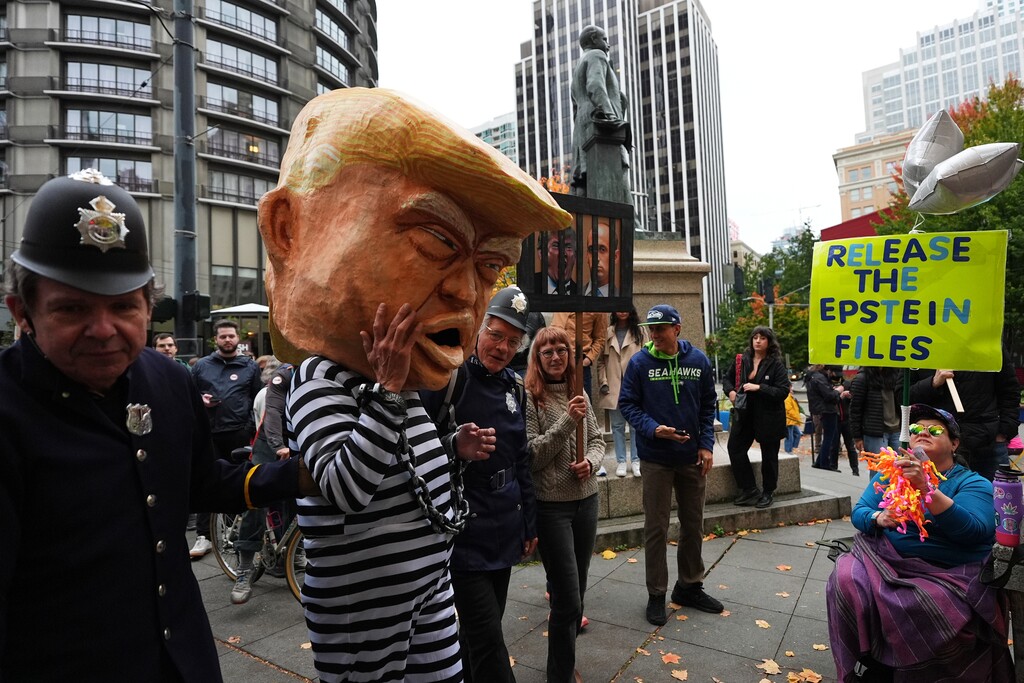 A protester dressed as President Donald Trump in chains and a prison jumpsuit looks at another holding a sign referencing the Epstein files as people take part in a "No Kings" protest Saturday, Oct. 18, 2025, in Seattle. (AP Photo/Lindsey Wasson)