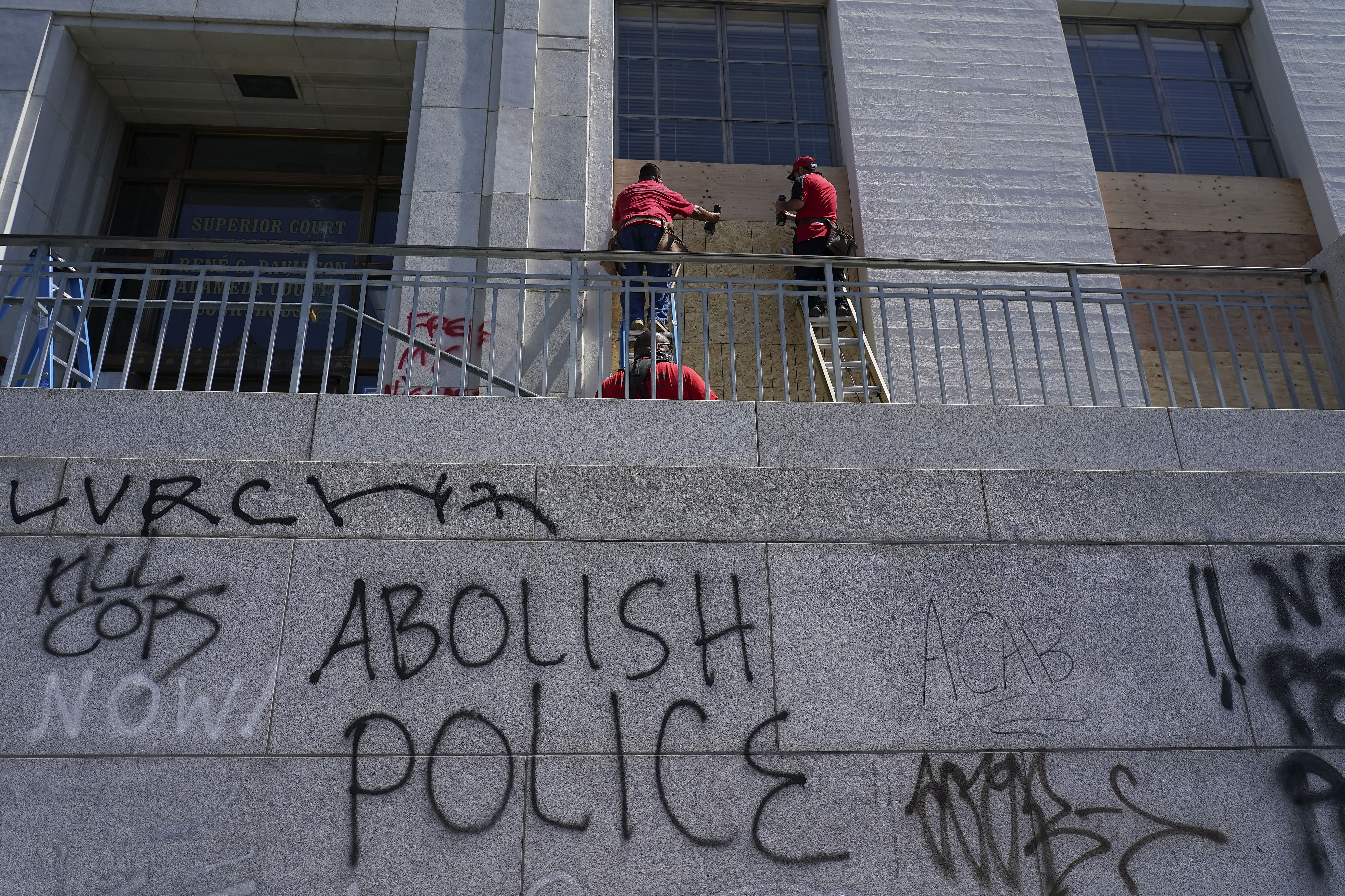 (Jeff Chiu | The Associated Press) Workers place wooden boards over windows as graffit is shown on walls at the Alameda County Courthouse in Oakland, Calif., Sunday, July 26, 2020. A protest Saturday night through the streets of downtown Oakland, California, in support of racial justice and police reform turned violent when "agitators" among the demonstrators set fire to a courthouse, vandalized a police station and shot fireworks at officers, authorities said.