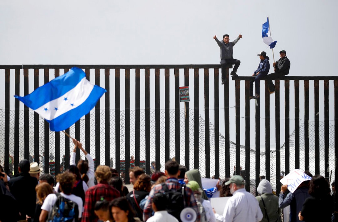 (Chris Carlson | The Associated Press)  Central American migrants sit on top of the border wall on the beach in San Diego during a gathering of migrants living on both sides of the border, Sunday, April 29, 2018.