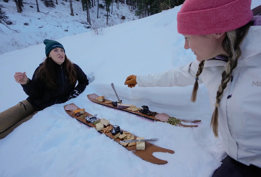 (Francisco Kjolseth | The Salt Lake Tribune) E.J. Elliott, left, and Kera Pezzuti, co-founders of Charcuteski, (pronounced “shar-koo-teh-ski”) enjoy nibbles after setting up a pair of ski blades in Summit Park to demonstrate their small Utah business venture on Monday, Dec. 11, 2023. The idea consists of going up into the mountains and eating a charcuterie spread off of skis or snowboards in the snow as a way to build community around snacking on the slopes.