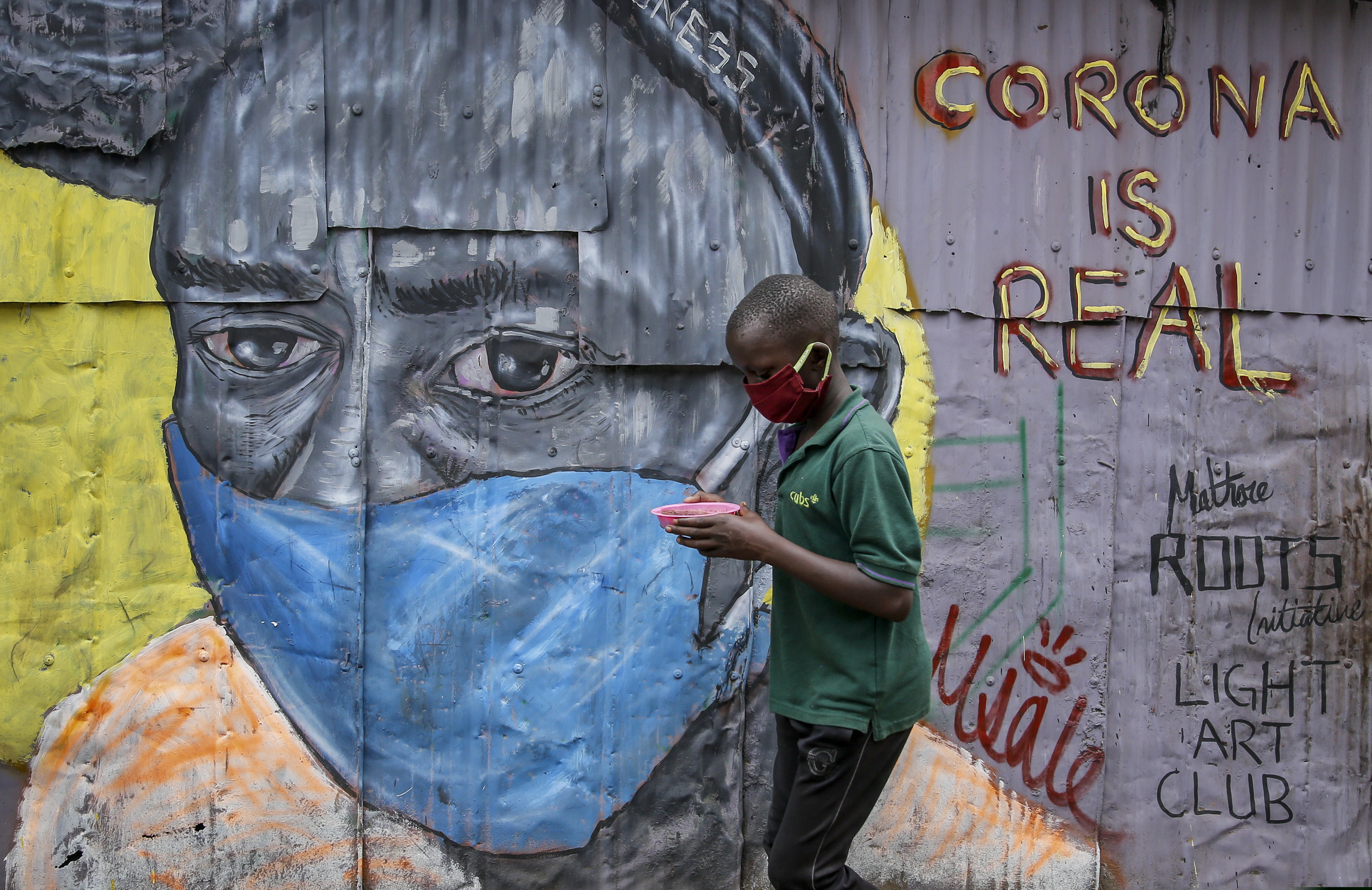 A boy wearing a face mask carries a small bowl of "githeri", or mixed beans and maize, for him to eat as he walks past an informational mural warning people about the risk of the new coronavirus, painted by graffiti artists from the Mathare Roots youth group, in the Mathare slum, or informal settlement, of Nairobi, Kenya Saturday, April 18, 2020. Africa now has more than 1,000 deaths from COVID-19, the Africa Centers for Disease Control and Prevention said Saturday, with 52 of the continent's 54 countries having reported cases. (AP Photo/Brian Inganga)