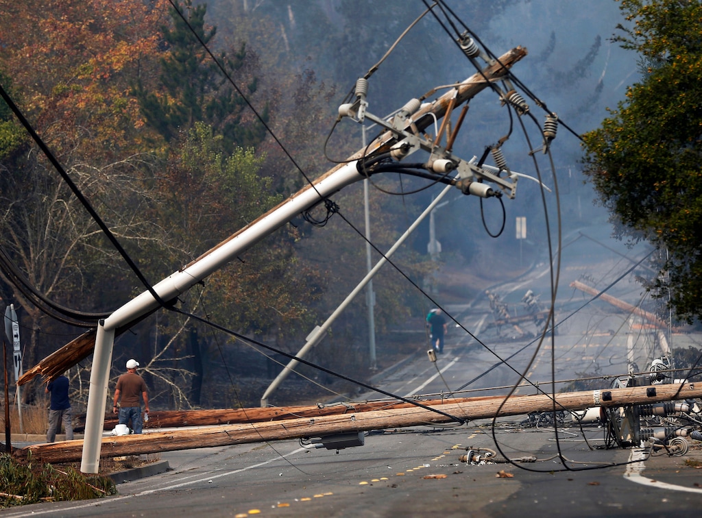 FILE - In this Tuesday, Oct. 10, 2017 file photo, people walk past a fallen transformer and downed power lines on Parker Hill Road in Santa Rosa, Calif. State fire officials blamed power lines coming into contact with trees for sparking four Northern California wildfires last October that incinerated more than 130 buildings. In a statement released Friday, May 25, 2018, officials indicated three of the fires could have been prevented if Pacific Gas & Electric Co. had made more efforts to keep trees clear of its power lines. PG&E says it is reviewing those conclusions. (Nhat V. Meyer/San Jose Mercury News via AP, File)