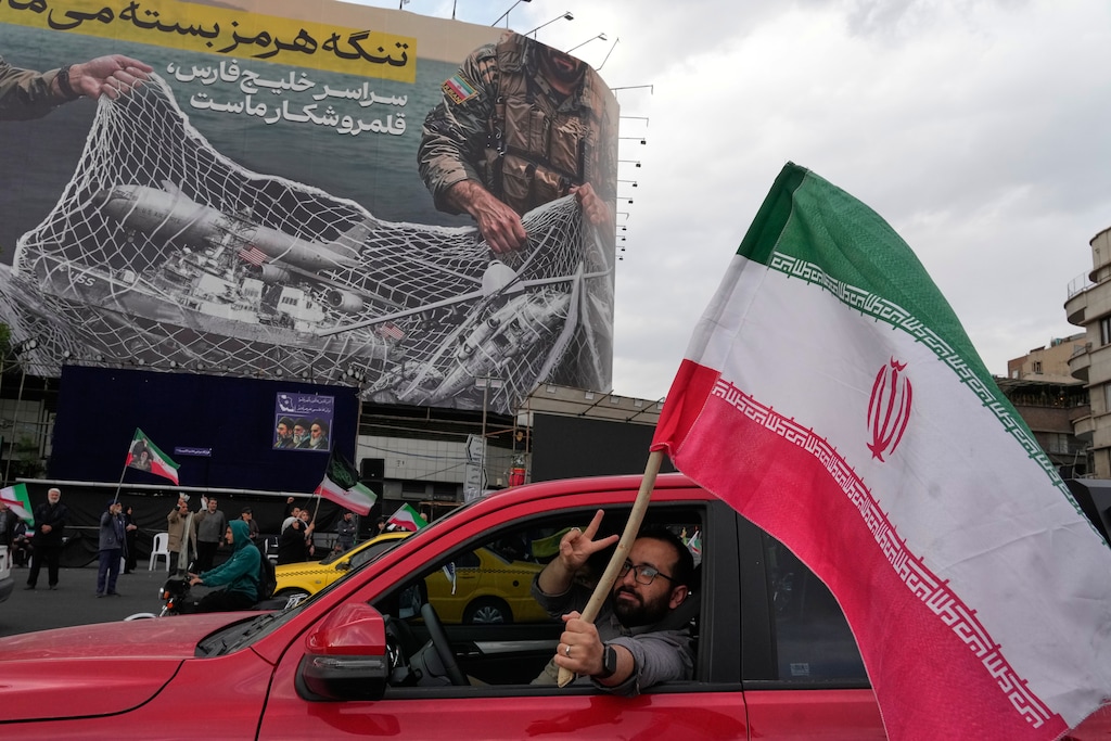 A man flashes a victory sign as he carries an Iranian flag in front of an anti-U.S. billboard depicting the American aircrafts in the Iranian armed forces fishing net with signs that read in Farsi: "The Strait of Hormuz will remain closed, The entire Persian Gulf is our hunting ground," at the Eqelab-e-Eslami, or Islamic Revolution Square in downtown Tehran, Iran, Monday, April 13, 2026. (AP Photo/Vahid Salemi)