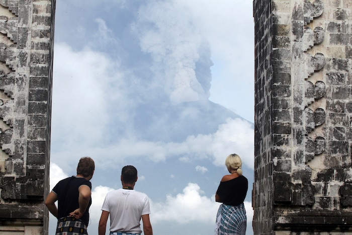 (Firdia Lisnawati | The Associated Press) Tourists watch the Mount Agung volcano erupting as they visit a temple in Karangasem, Indonesia, Monday, Nov. 27, 2017. Indonesia authorities raised the alert for the rumbling volcano to highest level on Monday and closed the international airport on tourist island of Bali stranding thousands of travelers.