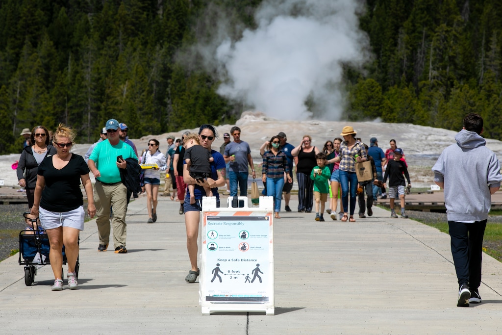 (Ryan Berry | Bozeman Daily Chronicle via AP) In this Monday, June 1, 2020 photo, visitors walk away from Old Faithful as a sign about COVID-19 safety sits in the sidewalk in Yellowstone National Park in Wyoming. Hundreds gathered at the Geyser for an afternoon eruption, and only a few dozen wore protective masks.