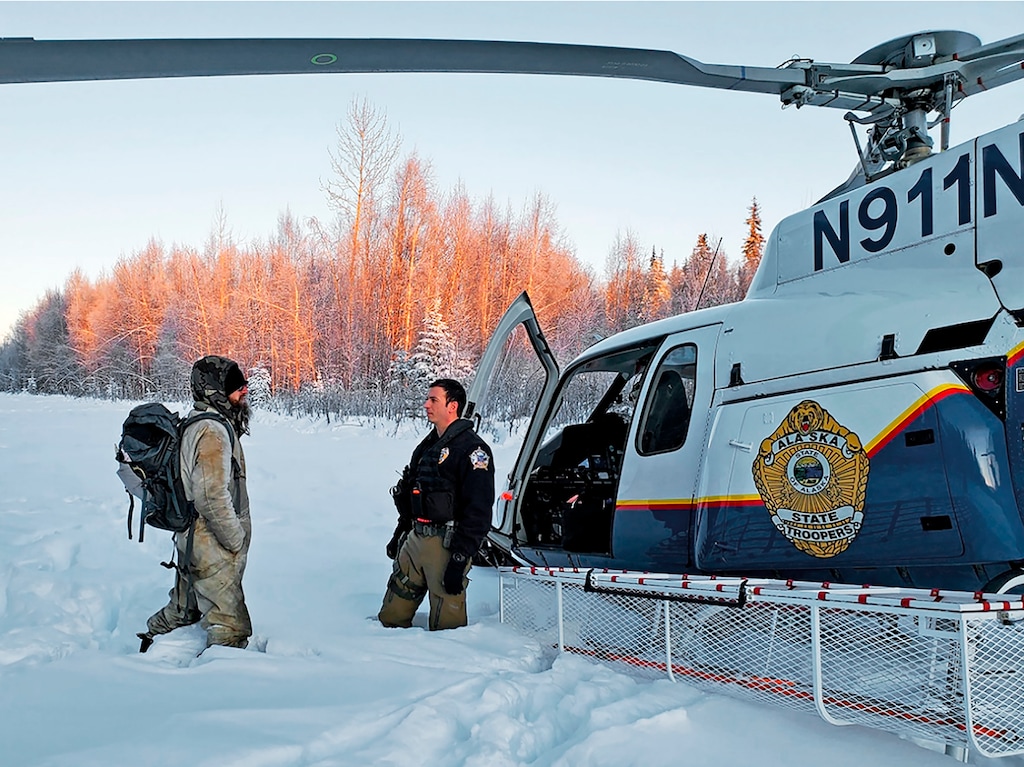 (Alaska State Troopers via AP) This Jan. 9, 2020 photo provided by Alaska State Troopers shows Tyson Steele, left, talking with Helo 3 Tactical Flight Officer Zac Johnson after Steele's rescue outside Susitna Valley, Alaska. Troopers rescued Tyson Steele, 30, who survived in a makeshift shelter after his remote cabin burned last month, after spotting him and an SOS sign in the snow.