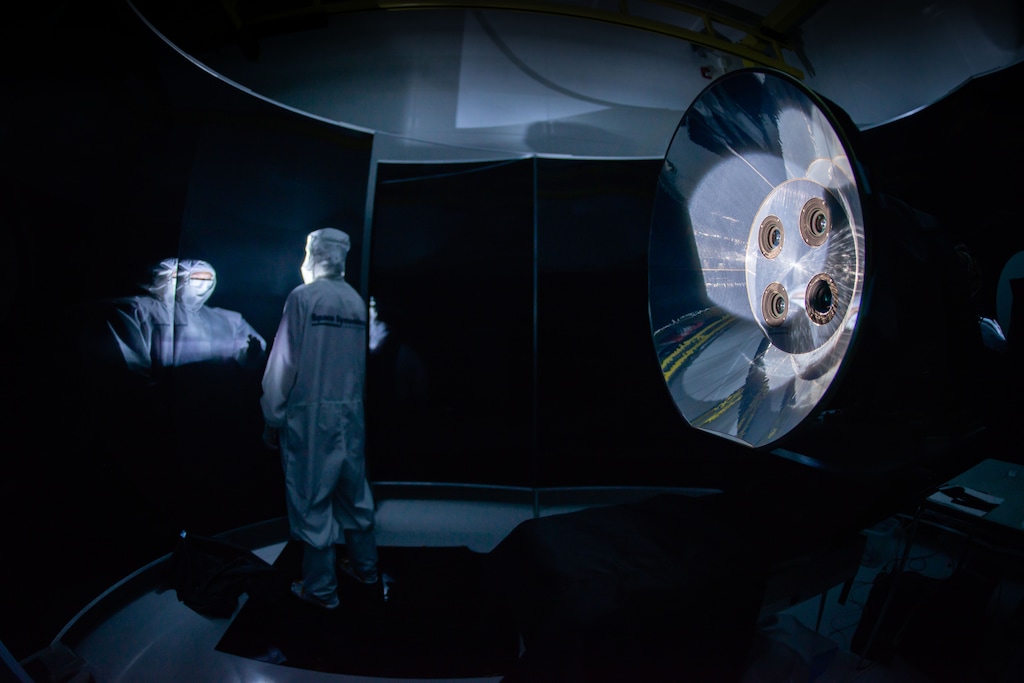 (Allison Bills | Space Dynamics Laboratory) A Space Dynamics Laboratory engineer standing next to the Atmospheric Waves Experiment (AWE) telescopes, with baffle, in a stray light test facility.