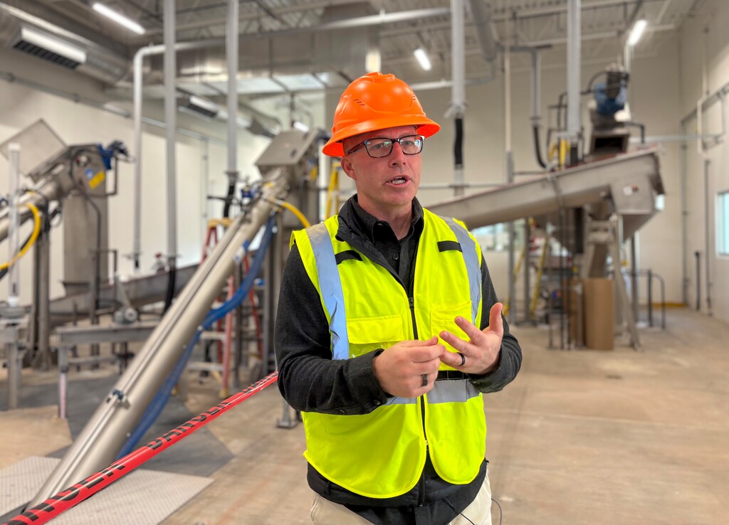 Mike Chandler, a white man stands in front of metal pipes. He wears a bright yellow vest and an orange hard hat.