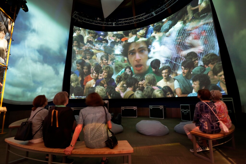 (Richard Drew | AP file photo) Visitors at the Museum at Bethel Woods, watch a video of the Woodstock Music and Art Fair, in Bethel, N.Y., on June 14, 2018. Aging baby boomers might blanch at the thought of archaeologists combing over the place that literally lent its name to their generation, as if it was a Civil War battle site. But Max Yasgur’s old farm about 80 miles north of New York City is already on the National Register of Historic Places.
