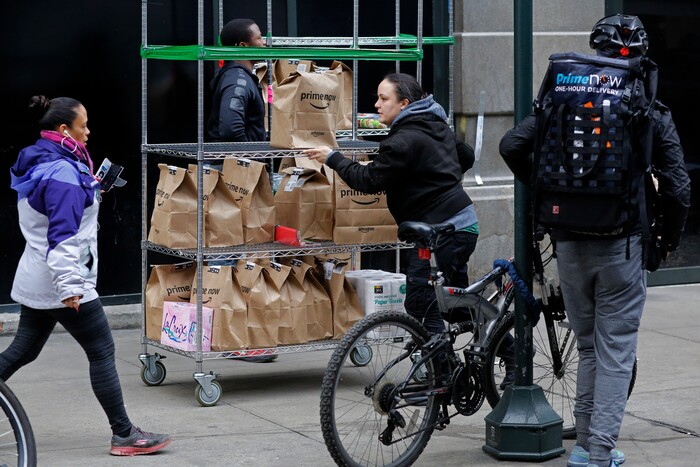 In this Wednesday, Dec. 20, 2017, photo, a clerk rolls a cart full of customer orders while a bicyclist with a backpack of orders prepares to leave the Amazon Prime warehouse, in New York. (AP Photo/Mark Lennihan)