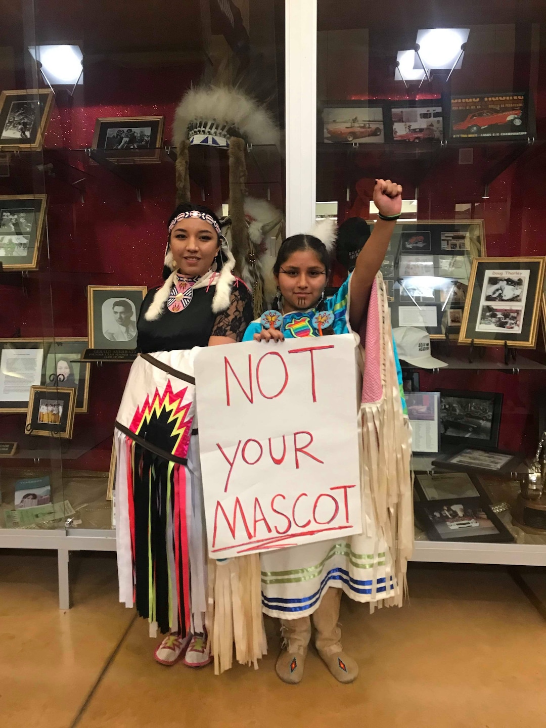(Photo courtesy of Marlee Kanosh) Cousins Jennilee Kanosh and Leia Shallenberger stand near a feather headdress in a trophy case at Cedar High School in Cedar City. CHS officials are considering dropping the school's controversial team name: The Redmen. While Kanosh and Shallenberger are not CHS students or alumni, they are members of the local Paiute Indian Tribe of Utah and attended a public hearing on Dec. 18, 2018, to advocate changing the mascot.
