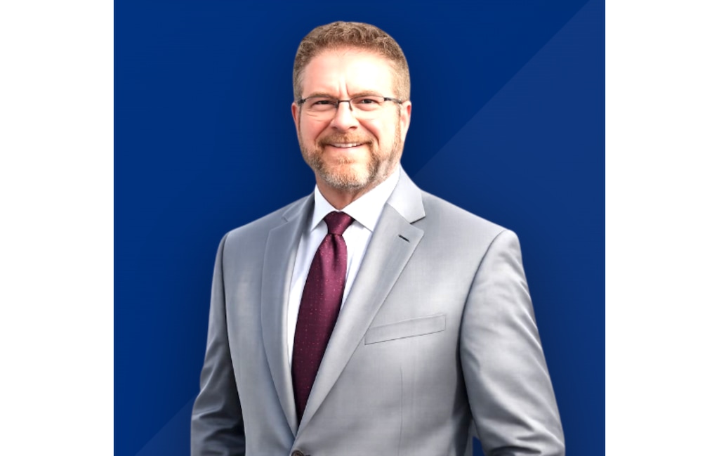 A man with a beard and short brown hair looks at the camera and smiles while wearing a gray suit and a maroon tie.