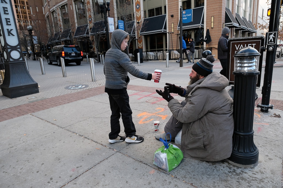 (Francisco Kjolseth  |  The Salt Lake Tribune) Ten-year-old Chase Hansen brings Chris Coons a coffee as he panhandles in downtown Salt Lake City on Monday, Dec. 16, 2019, in exchange for a little conversation. Chase and his father John have an organization called Project Empathy started when Chase was 6. The two of them spend time with people experiencing homelessness in an effort to connect with people on a personal level. On this occasion Chase learns that Coons used to be an electrician before losing much of his vision and how he loved to ski.