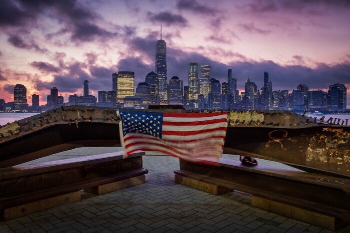 (J. David Ake | AP Photo) A U.S. flag hanging from a steel girder, damaged in the Sept. 11, 2001 attacks on the World Trade Center, blows in the breeze at a memorial in Jersey City, N.J., Sept. 11, 2019 as the sun rises behind One World Trade Center building and the re-developed area where the Twin Towers of World Trade Center once stood in New York City on the 18th anniversary of the attacks.