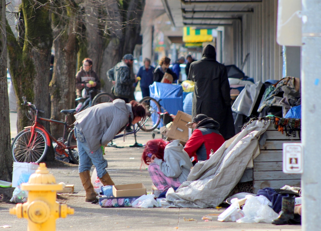 In this Tuesday, March 3, 2020 photo, homeless people crowd a sidewalk in downtown Salem, Ore., where they have set up a makeshift camp. Experts say that the homeless, who often have health and substance-abuse problems, are exposed to the elements and do not have easy access to hygiene, are more vulnerable to the coronavirus. Some cities are making provisions so the homeless who contract the virus have a place to recover without spreading the infection further. (AP Photo/Andrew Selsky)