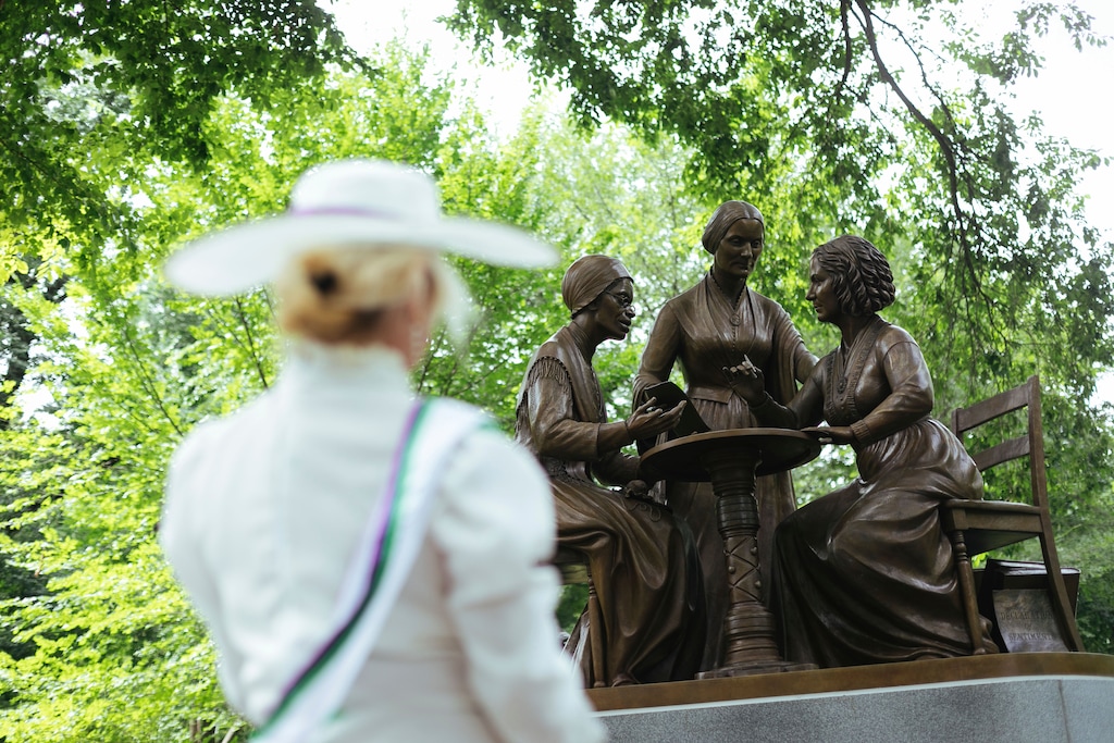 Mona Cahill, costumed as a women suffrage activist, stands in front of the Women's Rights Pioneers statue in Central Park Wednesday, Aug. 26, 2020, in New York. The statue, created by sculptor Meredith Bergmann and featuring Sojourner Truth, Susan B. Anthony and Elizabeth Cady Stanton, is the first monument in the park honoring any female historical figures. (AP Photo/Kevin Hagen)