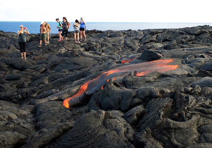 FILE - In this Monday, Aug. 8, 2016 file photo, visitors look at lava from Kilauea, an active volcano on Hawaii's Big Island, as it flows toward the ocean in Hawaii Volcanoes National Park near Kalapana, Hawaii. Hawaii residents and an organization representing federal workers filed a lawsuit against the Federal Aviation Administration on Wednesday, Oct. 4, 2017 seeking to force it to do something about tour helicopters buzzing their communities and national parks across the country. (AP Photo/Caleb Jones, File)
